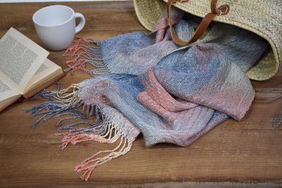 Handwoven and hand-painted scarf laying on a wooden table with a book and a mug.